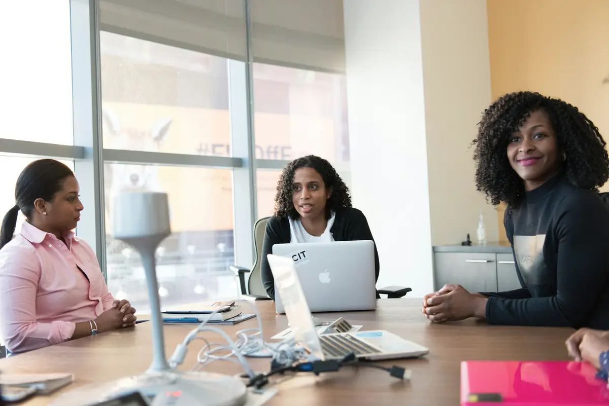 Three colleagues in discussion around a meeting table with laptops